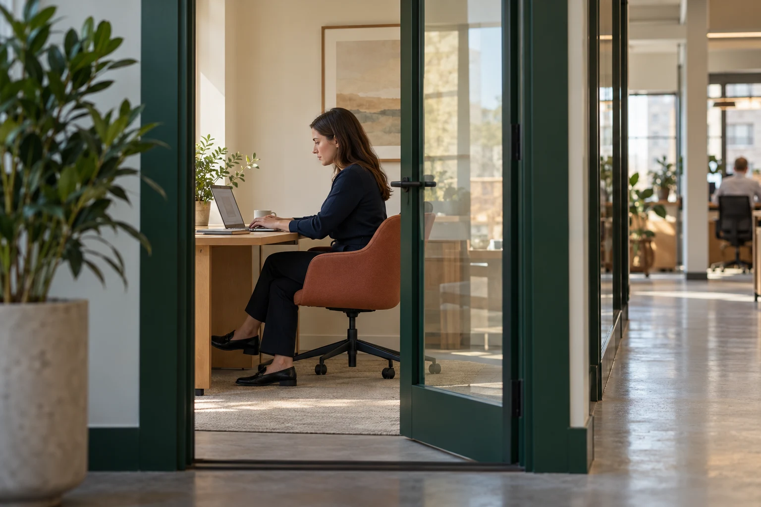 A woman works at a laptop in a glass-walled focus room with green doorframes and a terracotta chair.